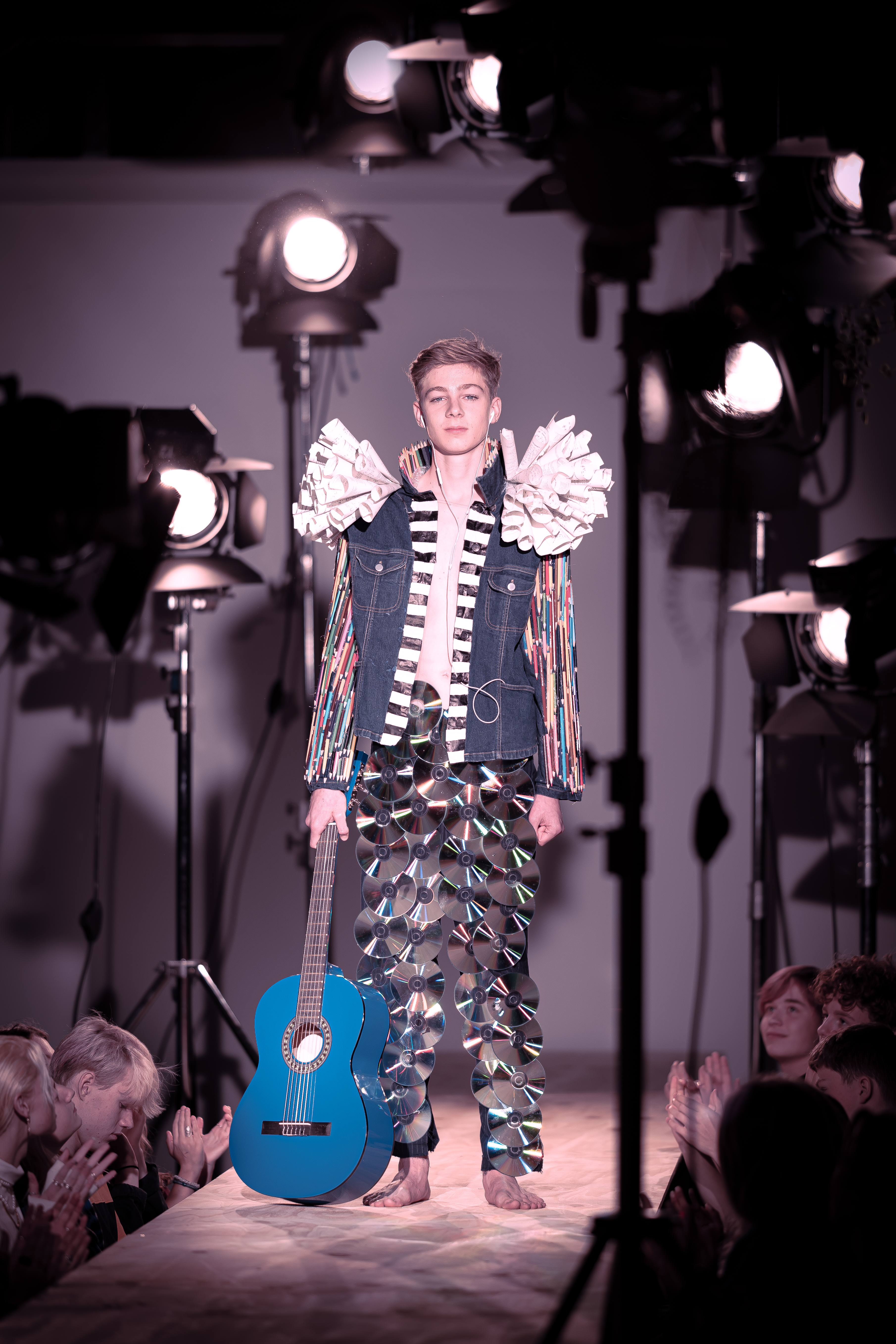 Ung model på catwalk i kreativt kostume af genbrugsmaterialer med blå guitar